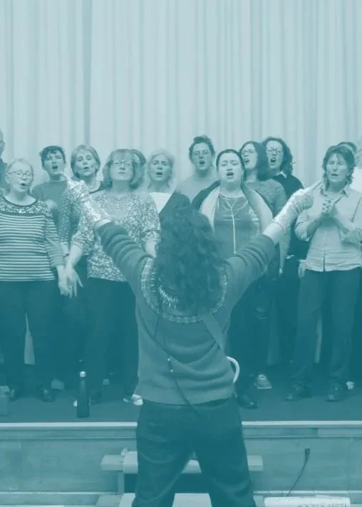 A blue tinted photo of the choir performing. Daisy is stood in front conducting with her back to the audience, arms stretched joyfully above her head.