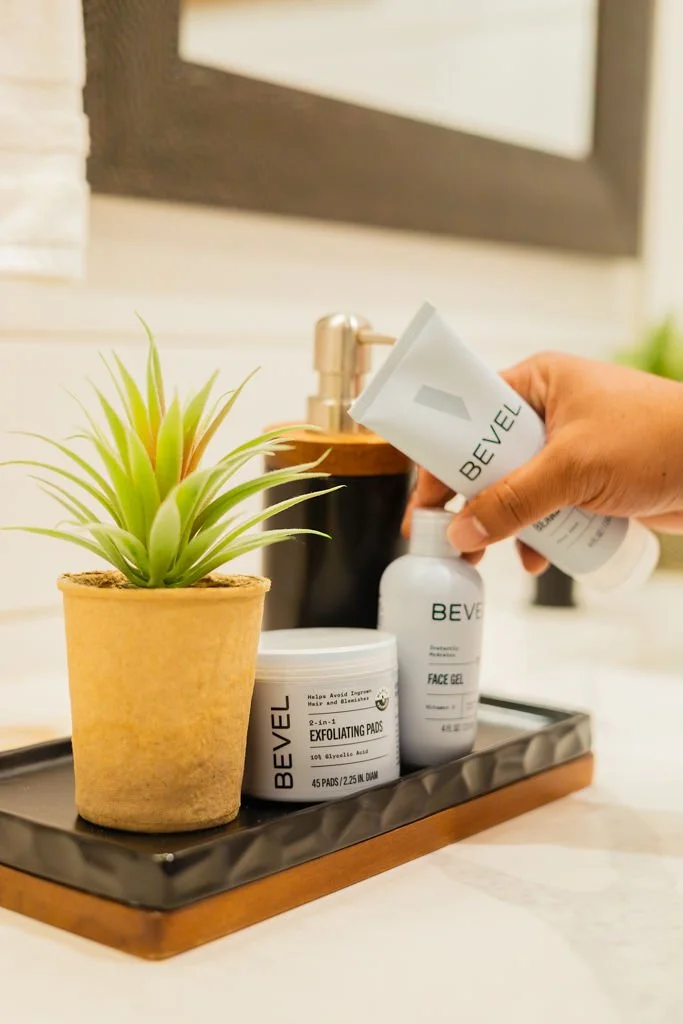 Person applying face gel near a potted green plant on a tray with exfoliating pads on a bathroom countertop