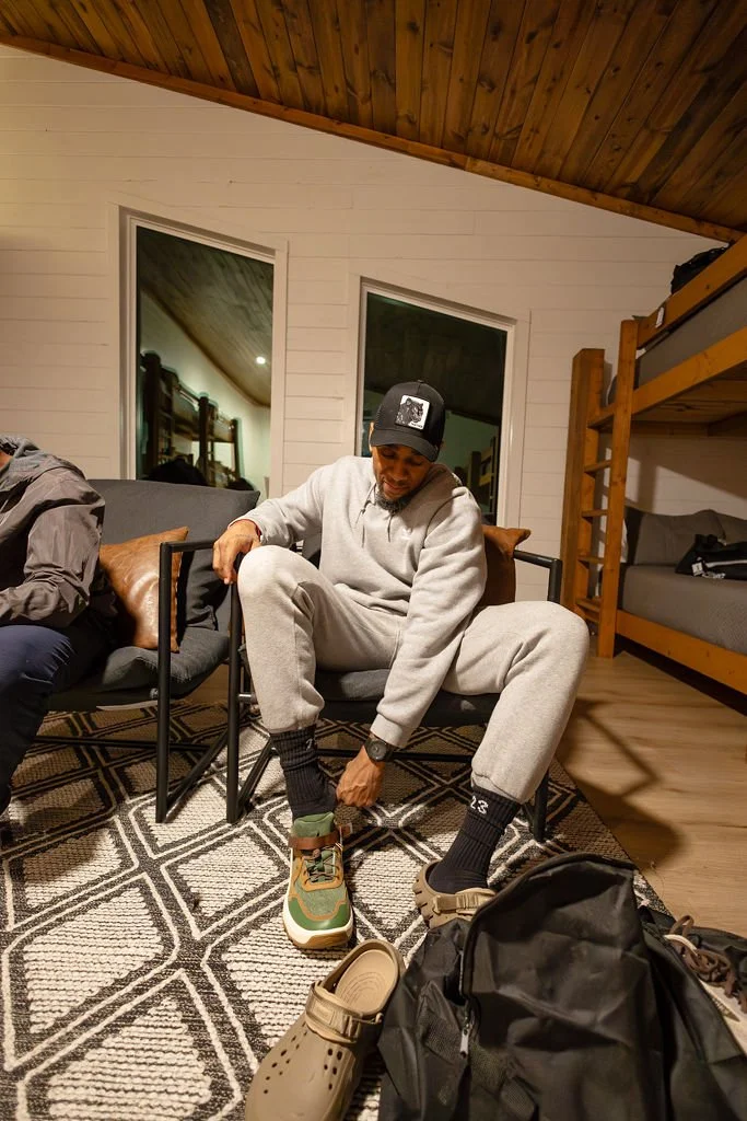 A man in a gray hoodie and sweatpants sitting on a chair, adjusting his sneaker while wearing a black cap. There is a black backpack and a tan shoe on the floor in front of him. The room has white walls, wooden ceiling, and a black patterned rug.