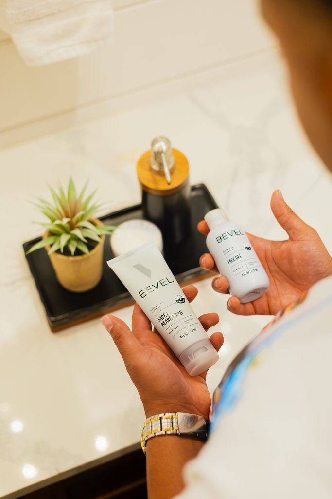 Person holding two bottles of BEVÉL face gel and face wash in front of a tray with a potted succulent plant, soap dispenser, and a container, on a white countertop.
