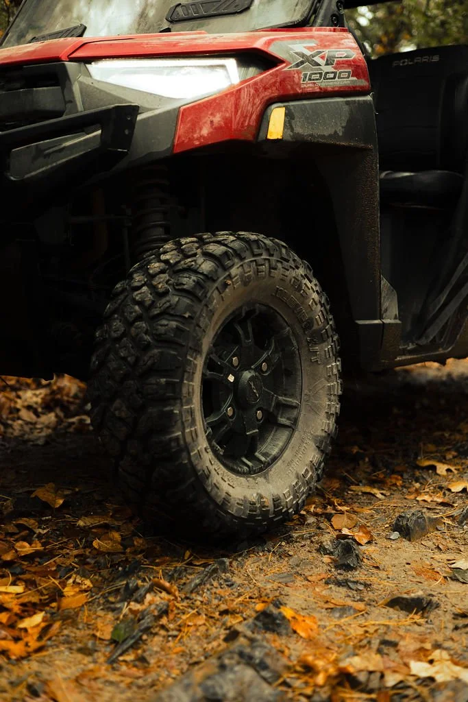 Close-up of a red Polaris XP 1000 side-by-side off-road vehicle with large, rugged tires on a dirt and leaf-covered ground.