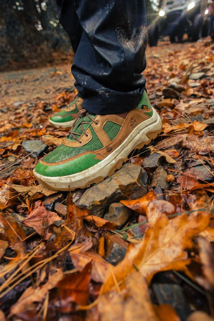 A person wearing green and brown trail shoes standing on a rocky, leaf-covered forest floor.