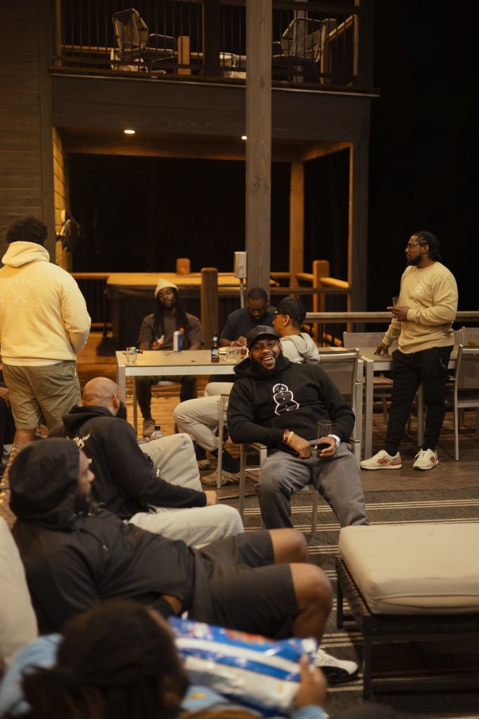 Group of men socializing indoors on a wooden deck, some seated on couches and chairs, others standing, with drinks present, in a cozy, warm-lit setting.