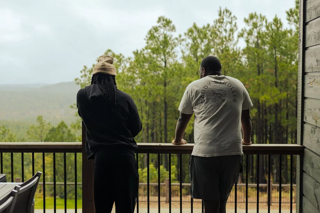Two people standing on a wooden balcony overlooking a green forested landscape.