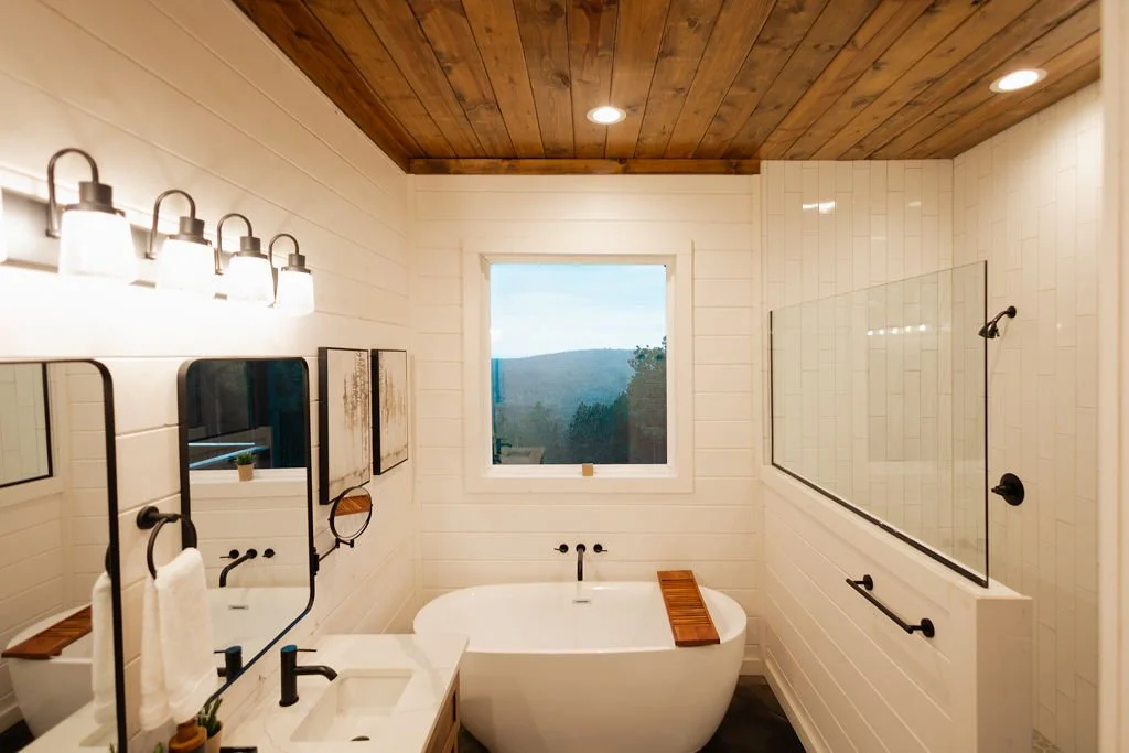 Modern bathroom with wooden ceiling, white shiplap walls, freestanding bathtub, large window with scenic view, walk-in shower with glass partition, black fixtures, and minimal decor.