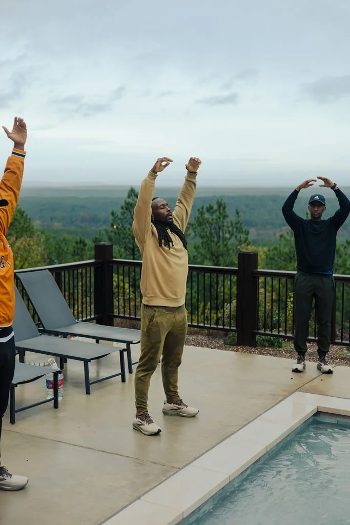 Three men are standing on a deck, mid-stretch, with a scenic view of trees and a cloudy sky in the background. They are near a pool and outdoor lounge chairs.