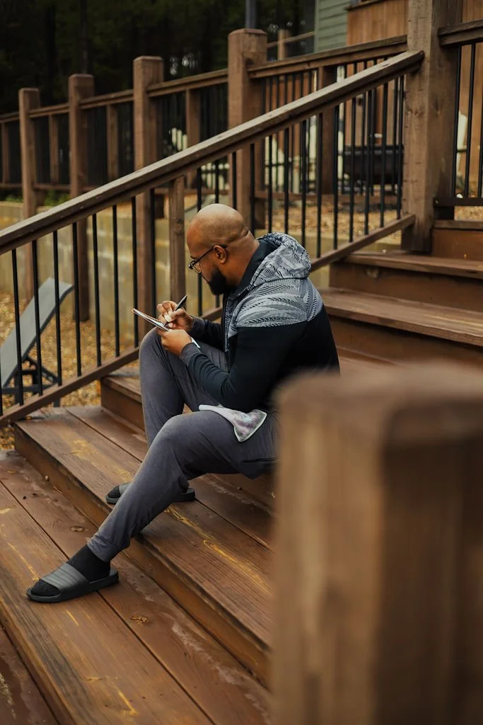 A man sitting on wooden stairs outside, using a spiral notebook and pen, wearing glasses, a black and gray jacket, gray sweatpants, and black slides.