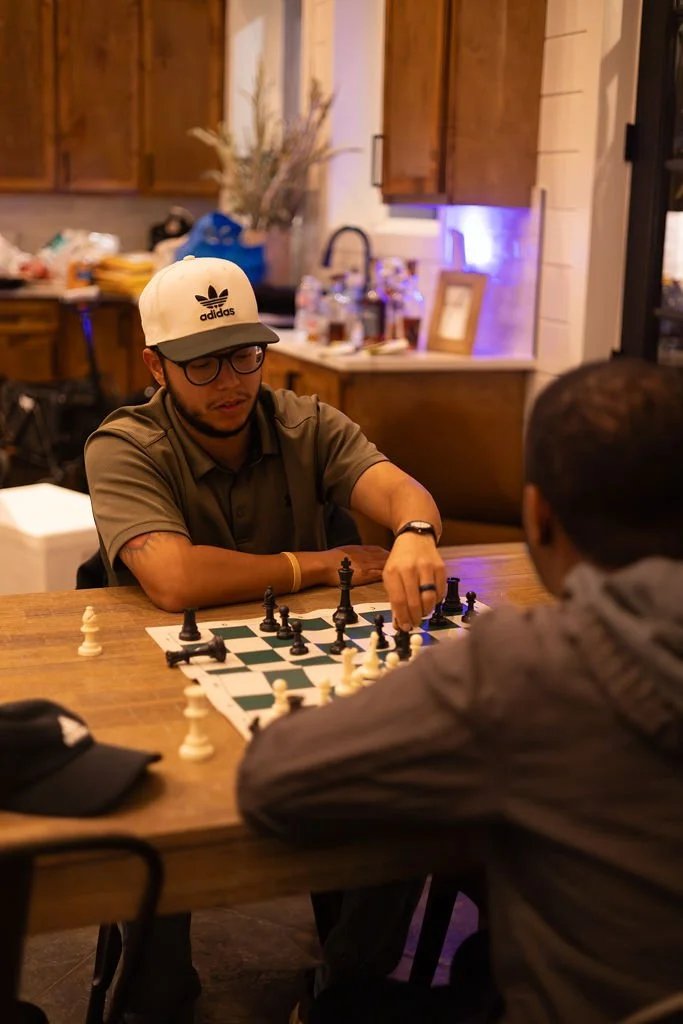 Two young men playing chess at a wooden table in a cozy kitchen. One is wearing glasses, a beige Adidas cap, and a brown shirt, while the other has dark hair and a hoodie. The kitchen has wooden cabinets, a sink, and various kitchen items in the back