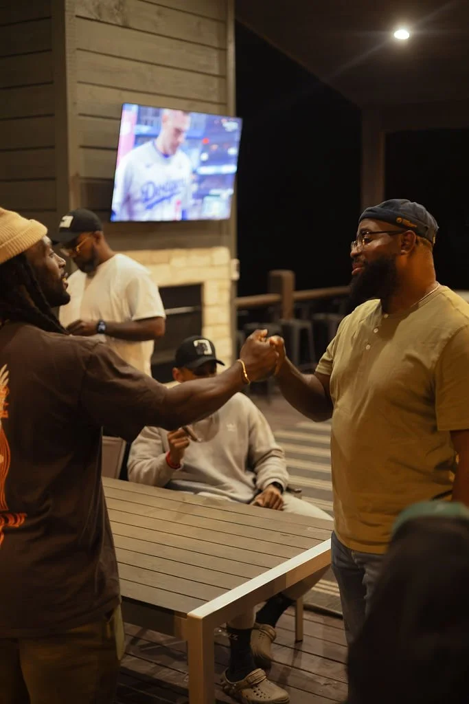 Two men shaking hands in a friendly manner at a restaurant or bar, with a group of people sitting at a table and a television showing a sports game in the background.