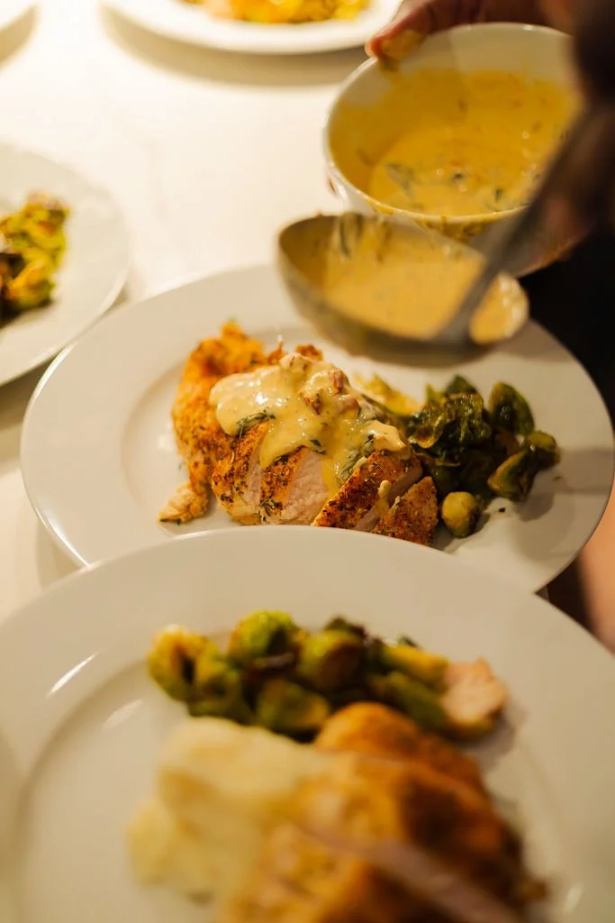 Person pouring creamy sauce over breaded chicken with vegetables on a white plate.
