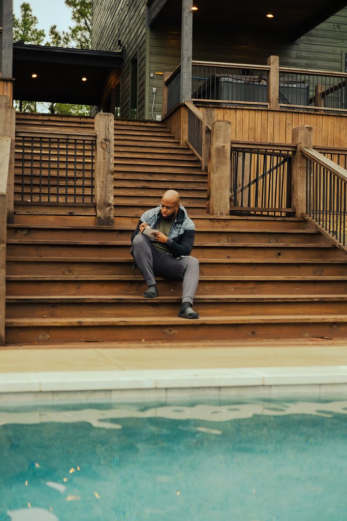 A man sits on wooden steps by a pool, looking at his phone. The steps lead up to a house with a deck.