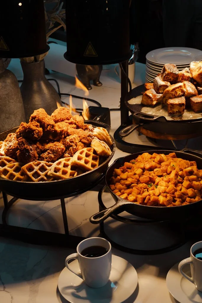 Fried chicken and waffles, sweet potatoes, and breakfast pastries on a buffet table, with cups of coffee in front.