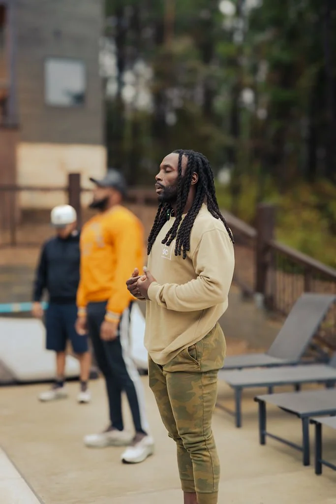 A man with long dreadlocks standing outdoors, wearing a beige hoodie and camouflage pants, with two other people blurred in the background.