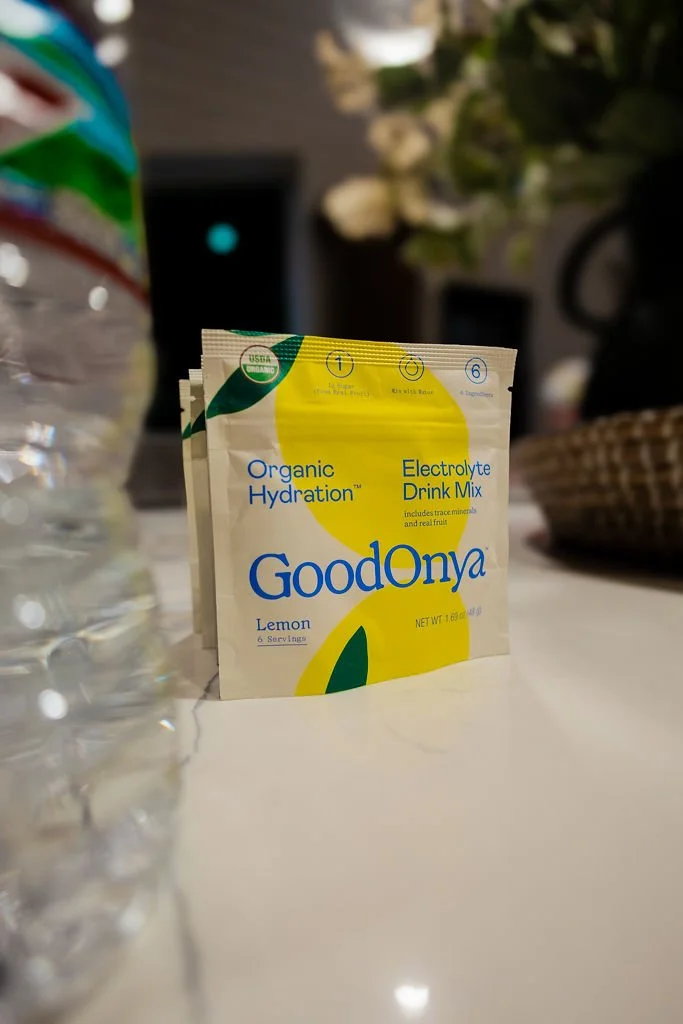 A packet of GoodOnya Organic Hydration Lemon electrolyte drink mix on a table, with a blurred background including a water bottle, a laptop, and a basket.