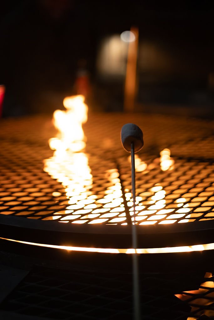 A marshmallow on a stick roasting over an open flame on a metal fire pit at night.