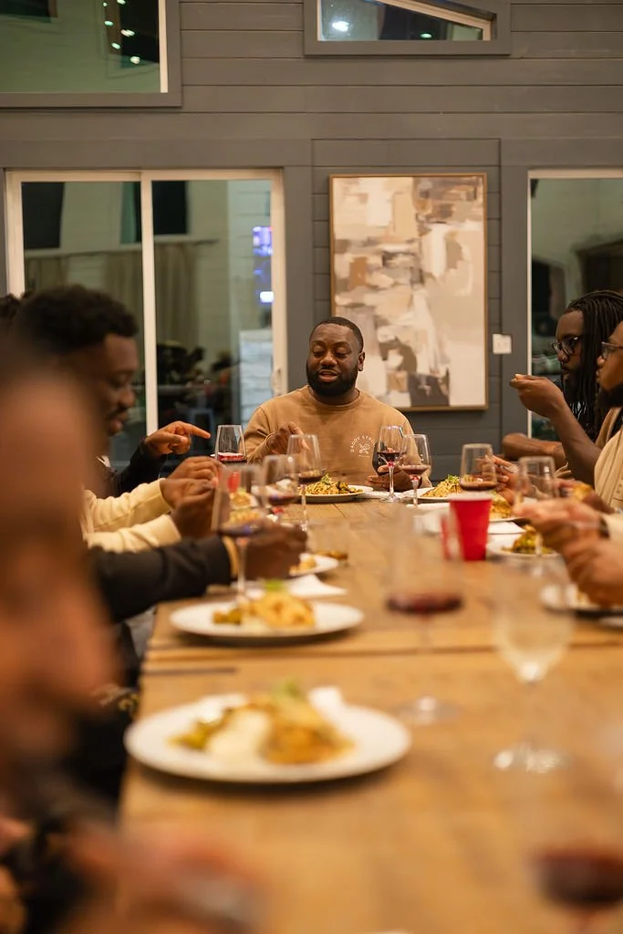 A group of people gathered around a long wooden dining table, enjoying a meal together in a cozy indoor setting, with glasses of red wine and plates of food.