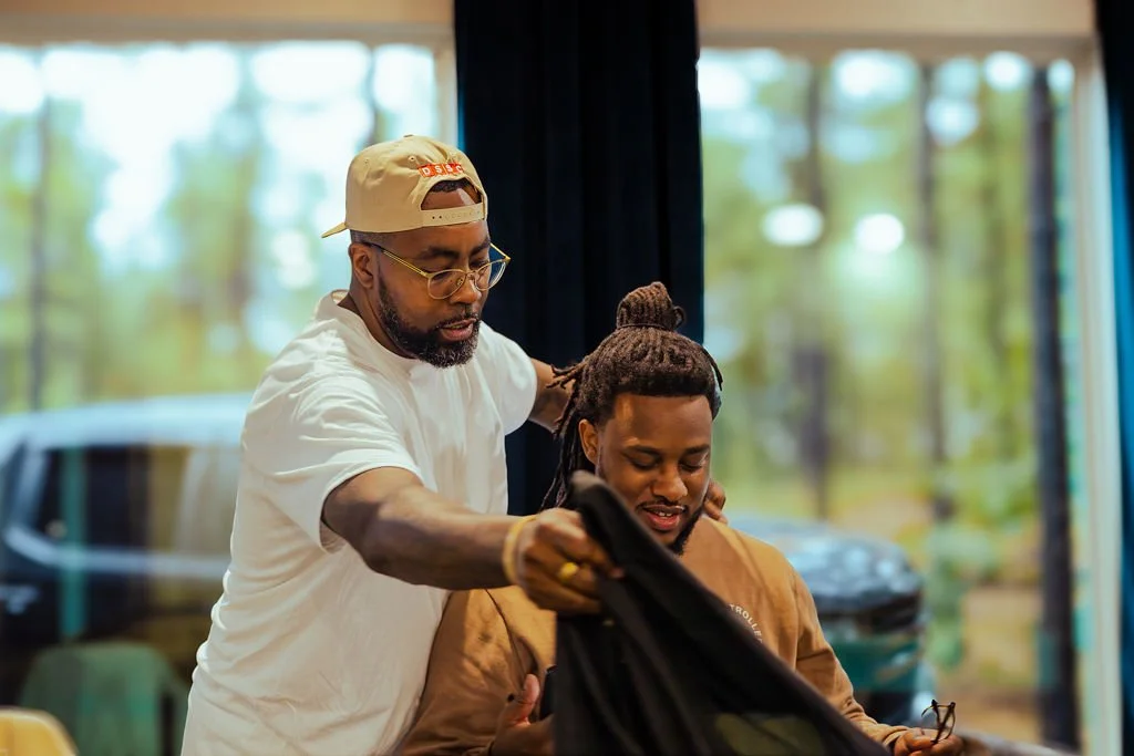 A man with glasses and a cap helps a seated man with dreadlocks put on a hoodie inside a room with large windows and a blurry outdoors background.