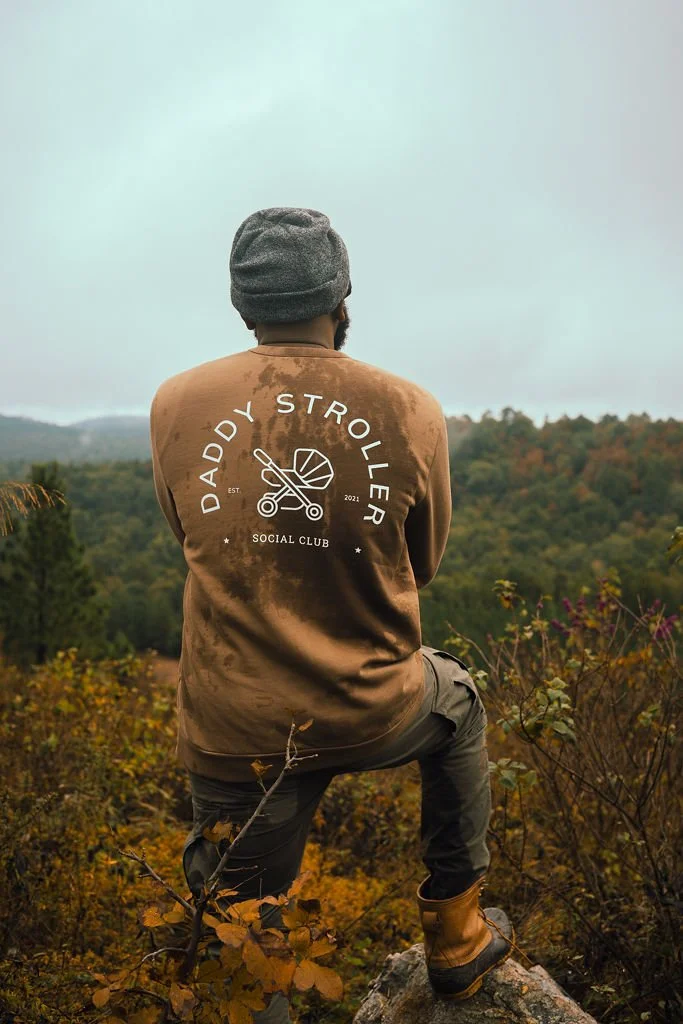 A man in outdoor clothing looking at a scenic landscape with trees and hills, wearing a brown jacket with 'Daddy Stroller' logo on the back.
