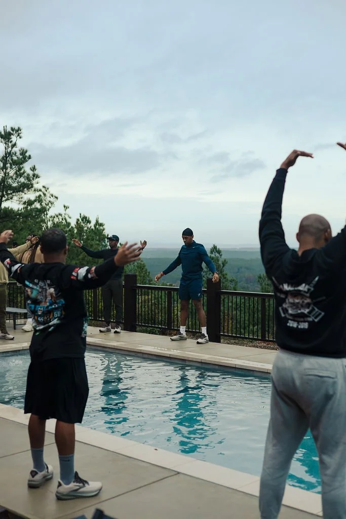 Group of people standing around an outdoor pool, some with arms raised, overlooking a scenic landscape with trees and cloudy sky.