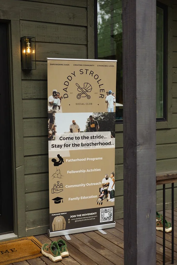 Banner for Daddy Stroller Social Club on a porch, promoting fatherhood programs, fellowship activities, community outreach, and family education, with a QR code and a pair of green and beige sneakers on a doormat.