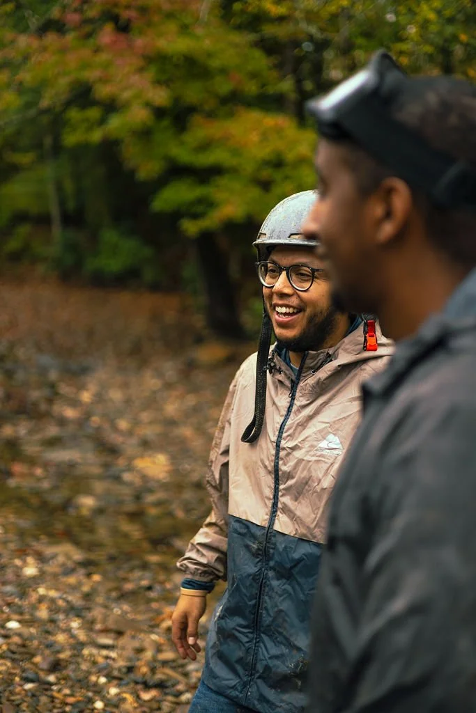 Two men outdoors in a wooded area, smiling and wearing jackets and helmet, surrounded by fall foliage.