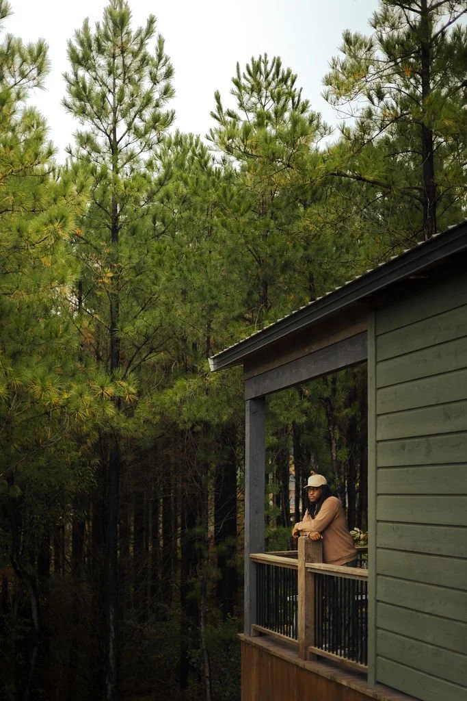 A person leaning on a balcony railing outside a green house, surrounded by tall pine trees.