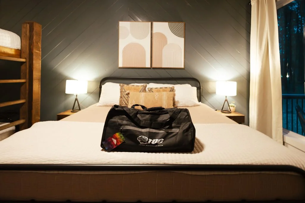 A hotel room with a large bed, two nightstands with lamps, a cozy pillow arrangement, a colorful abstract wall art, a wooden bunk bed to the left, and a balcony door to the right with curtains, with a black sports bag on the bed.