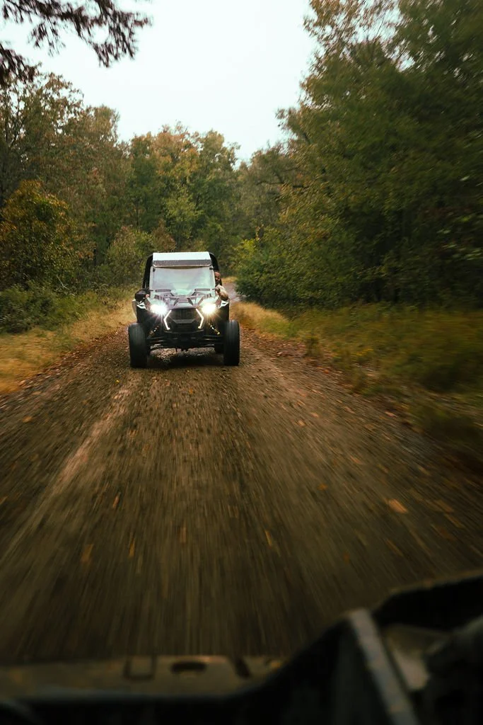 An off-road vehicle driving on a dirt trail surrounded by dense trees during daytime.