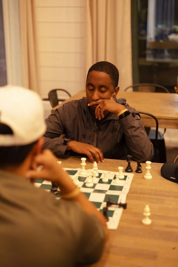 Two young men playing chess at a wooden table indoors, with a serious expression. One is wearing a gray jacket and gold wristwatch, the other has a white hat and is partially visible.