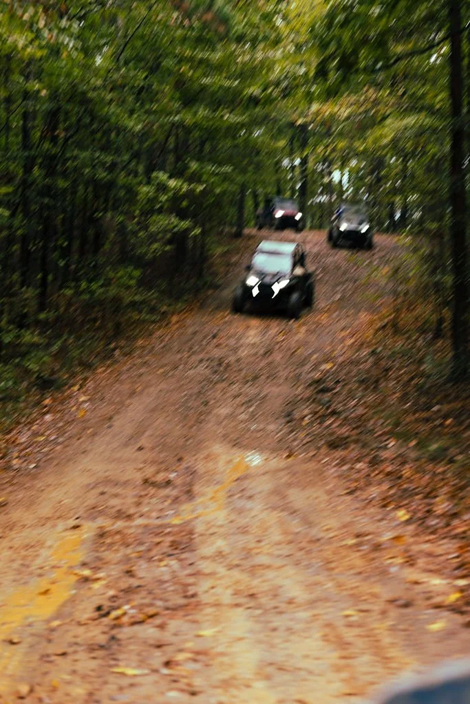 Three black off-road vehicles driving on a dirt path through a dense forest with green trees.