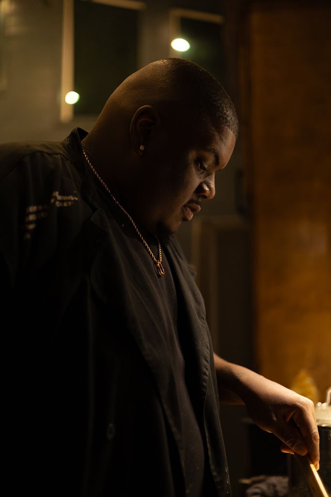 A man with a shaved head and earrings, wearing a black jacket and a silver chain, is looking down at something out of view in a dimly lit room with warm lighting.