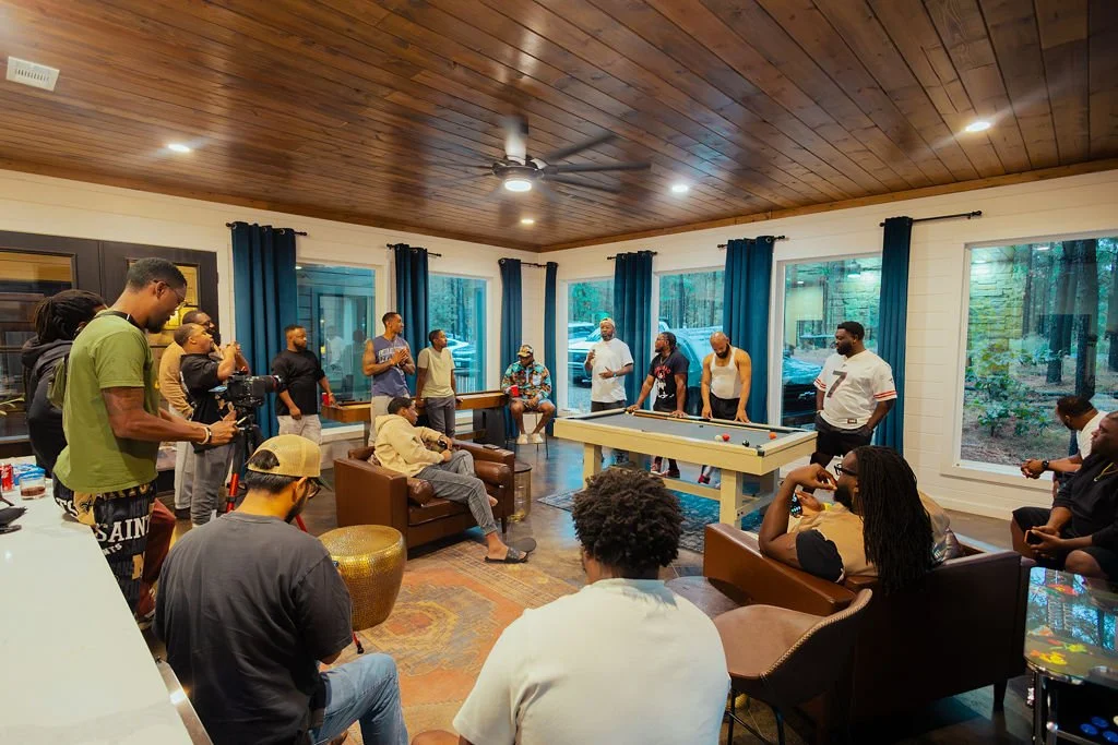 Group of people gathered in a living room watching a game of pool, with some taking photos and videos. The room has wooden ceilings, large windows with blue curtains, and modern furniture.