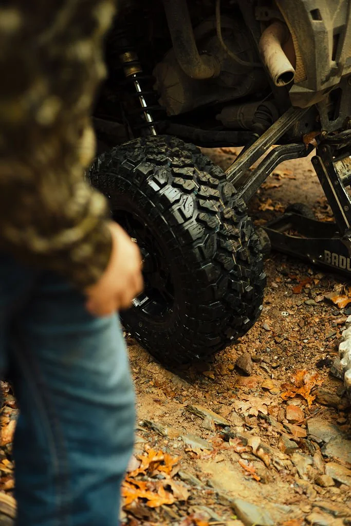 Close-up of a rugged off-road tire on a vehicle, with a person in the foreground outdoors on a dirt trail with leaves.
