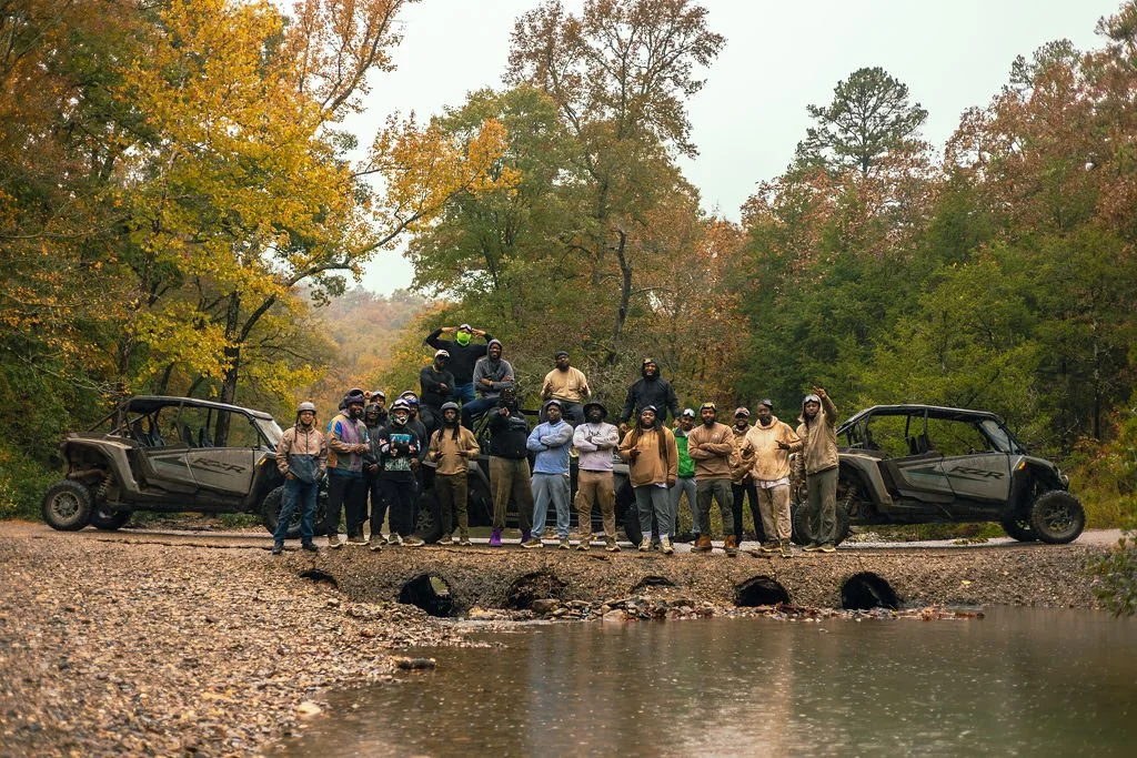 Group of people standing on a dirt road beside a creek, surrounded by trees with autumn foliage, with two off-road vehicles on either side.