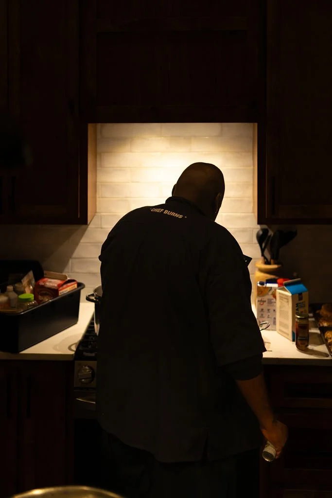 A man wearing a chef's jacket labeled 'Chef Burns' Burns Kitchen preparing food in a kitchen under warm lighting, with various ingredients and kitchen utensils on the countertop.