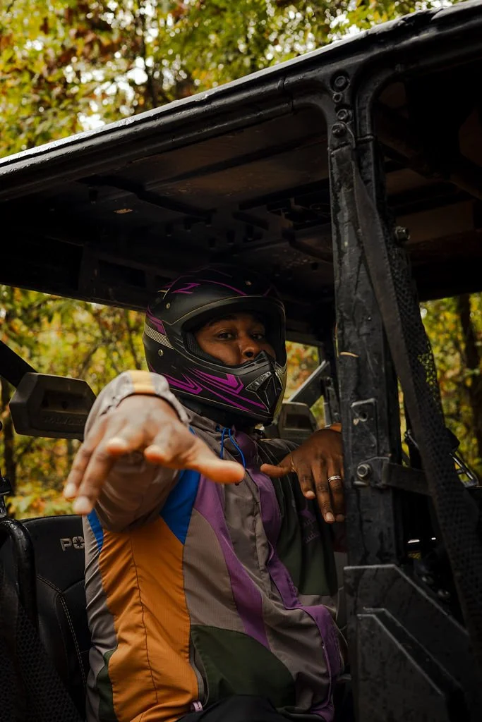 Person wearing a motorcycle helmet and colorful jacket sitting in a vehicle, pointing forward with one hand, outdoors with trees in the background.