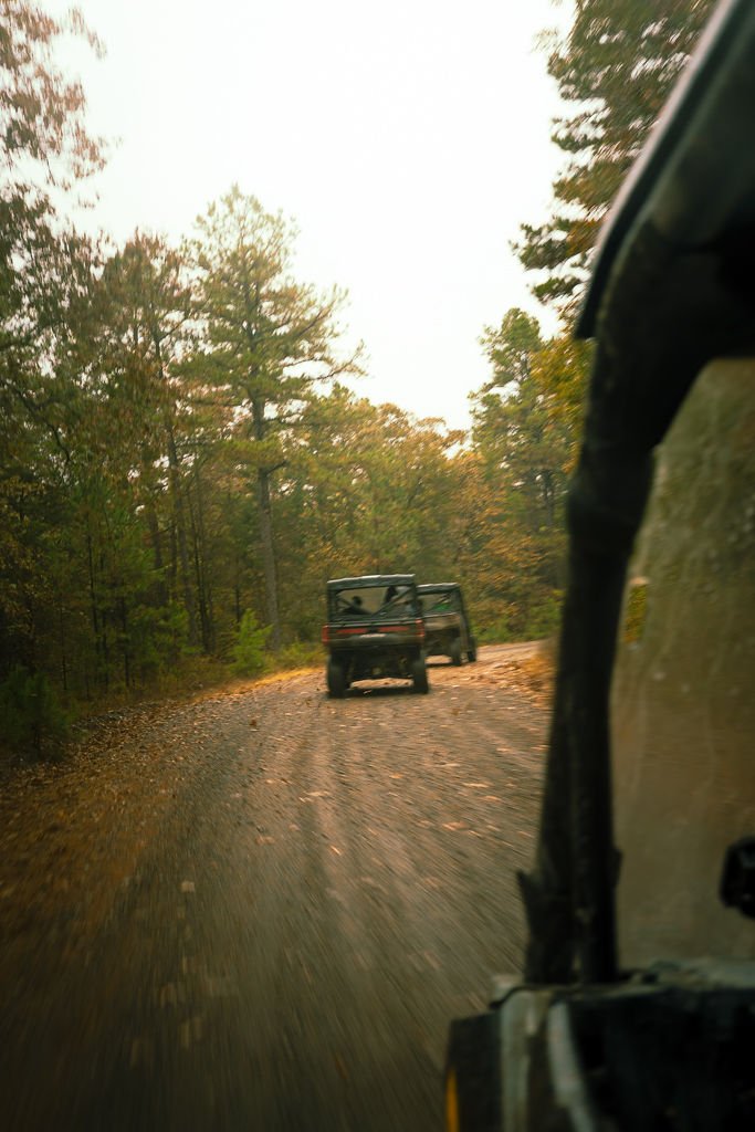 View from an off-road vehicle on a dirt trail surrounded by trees with autumn foliage, with another vehicle ahead on the trail.
