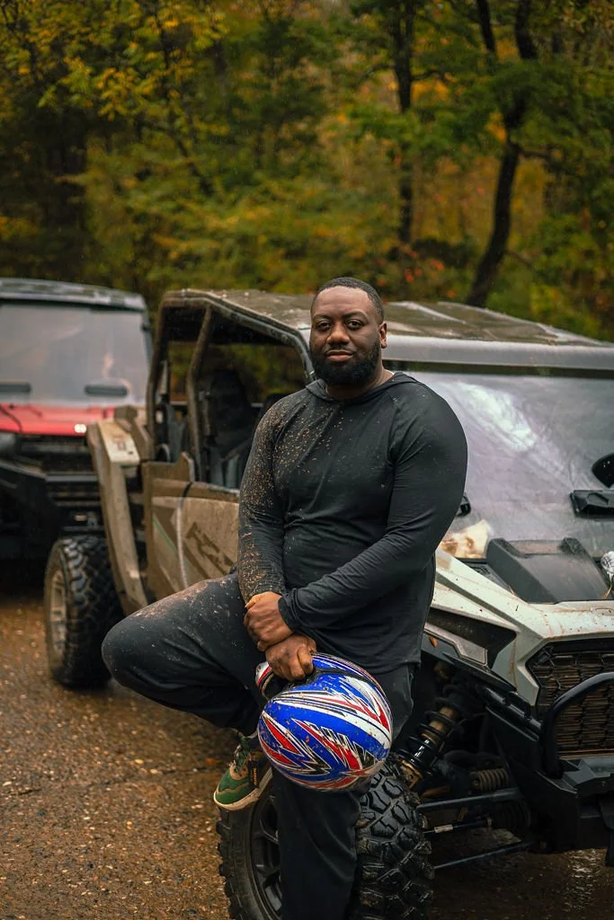 A man holding a motorcycle helmet stands in front of off-road vehicles in a wooded area during autumn.