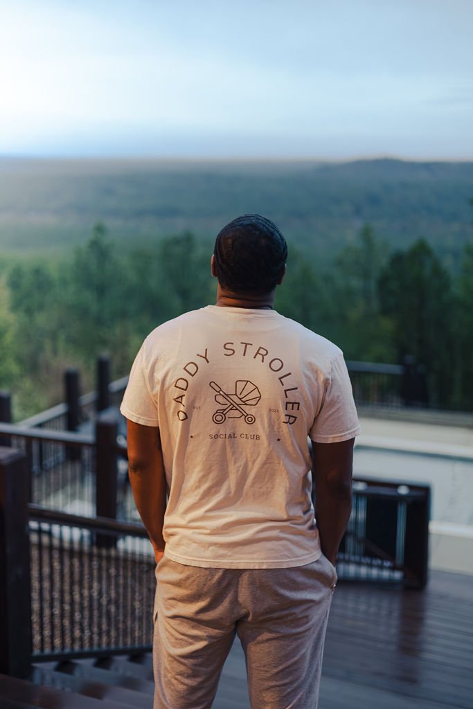 A man standing on a balcony overlooking a lush green landscape with hills and trees in the distance at sunset or sunrise.