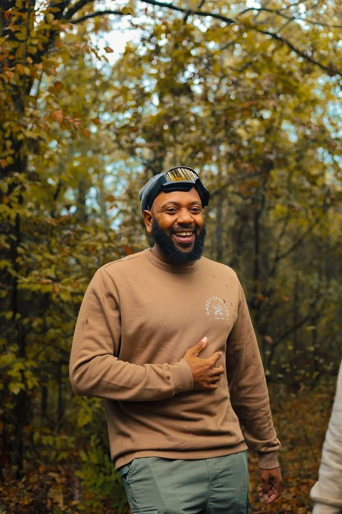 A man outdoors in a wooded area during fall, smiling, wearing a brown sweatshirt and gray pants, with a beanie and goggles on his head.