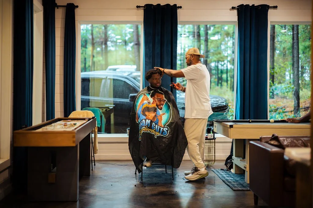 A man getting a haircut at a barbershop inside a house, with large windows showing a wooded outdoor scene.