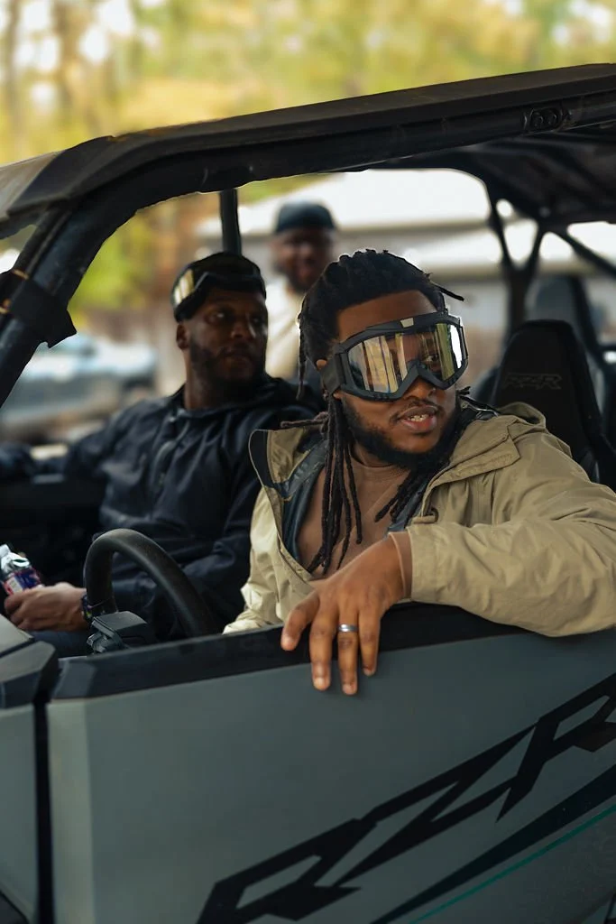 Three men in a side-by-side off-road vehicle. The man in the front wears goggles, a beige jacket, and has dreadlocks. The man in the back seats wears a black jacket, and the third man is blurred in the background.