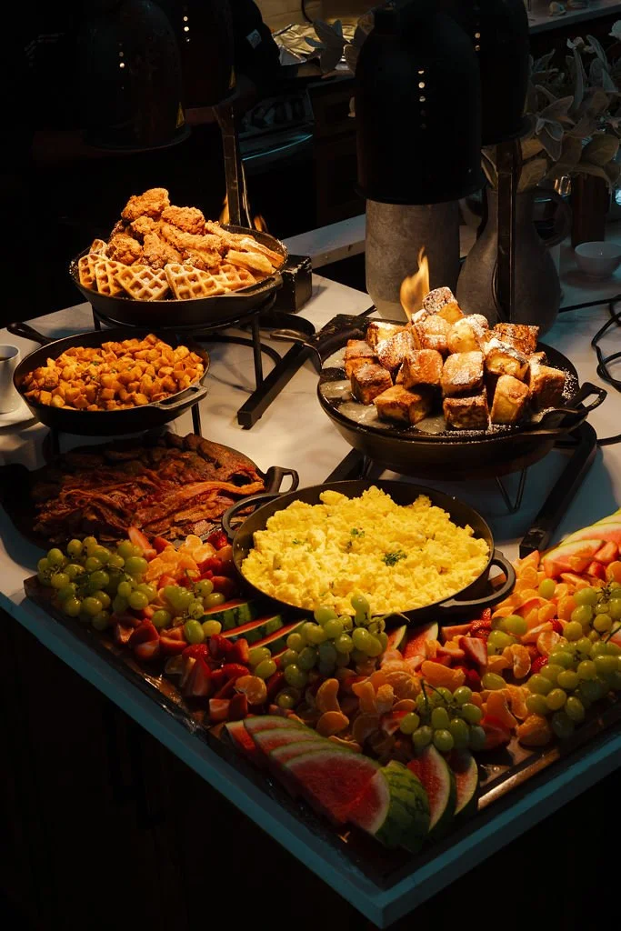 A buffet table with fried chicken, waffles, scrambled eggs, bacon, fresh fruit, and various breakfast items.