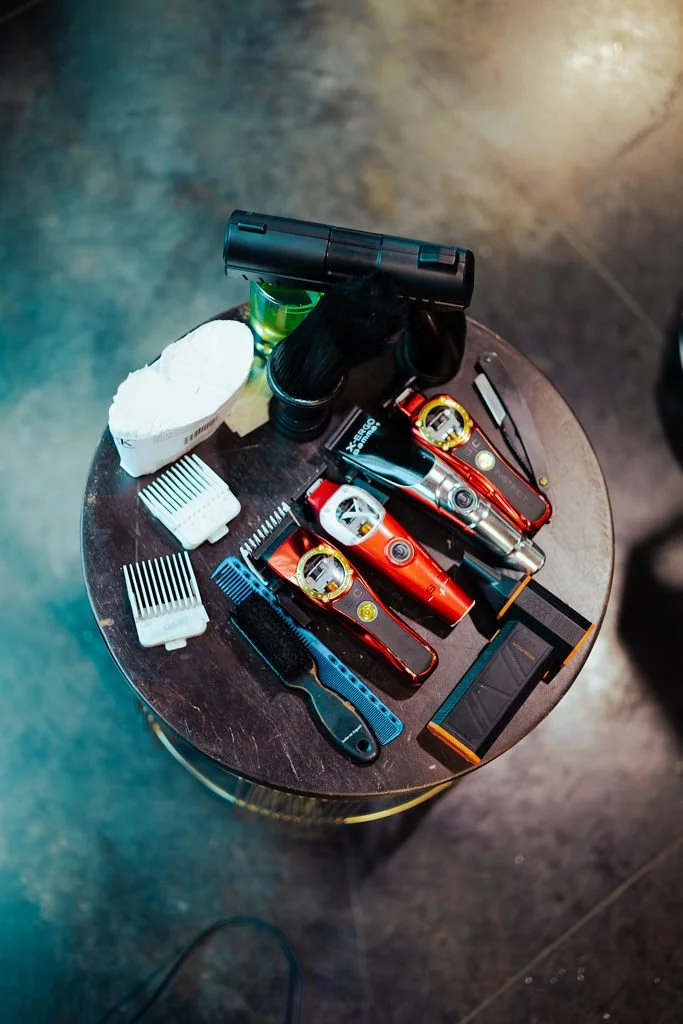 Various hair grooming tools including electric razors, trimmers, guards, scissors, a comb, a spray bottle, a towel, and other grooming accessories on a dark round table.