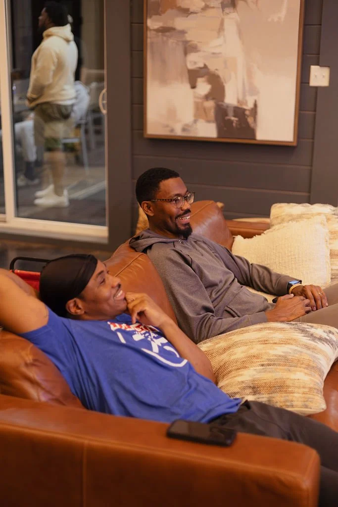 Two men sitting on a brown leather couch, smiling and laughing, with a large abstract painting on the wall behind them. One is wearing glasses and a gray hoodie, the other is in a blue shirt and black head covering. A phone is resting on the armrest 