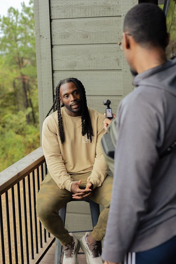 A man with long dreadlocks sitting on a porch being interviewed by another man holding a recording device.