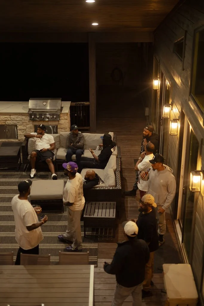Group of people socializing on a patio at night, some sitting on a sofa, others standing, with a grill and brick wall in the background.
