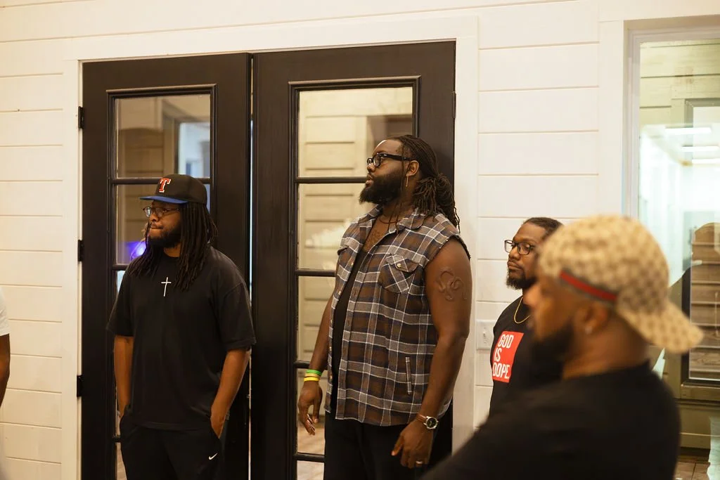 Four men standing indoors in front of a black door with glass panels, with white paneled walls, and a window, appearing to be having a conversation or listening attentively.
