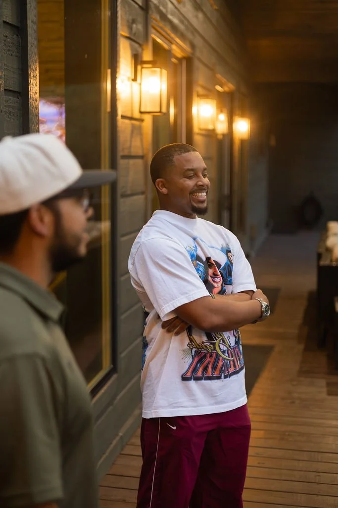 Two men indoors at a social gathering, one smiling with arms crossed, wearing a graphic T-shirt and maroon pants, and the other blurred in the foreground wearing a hat and glasses.