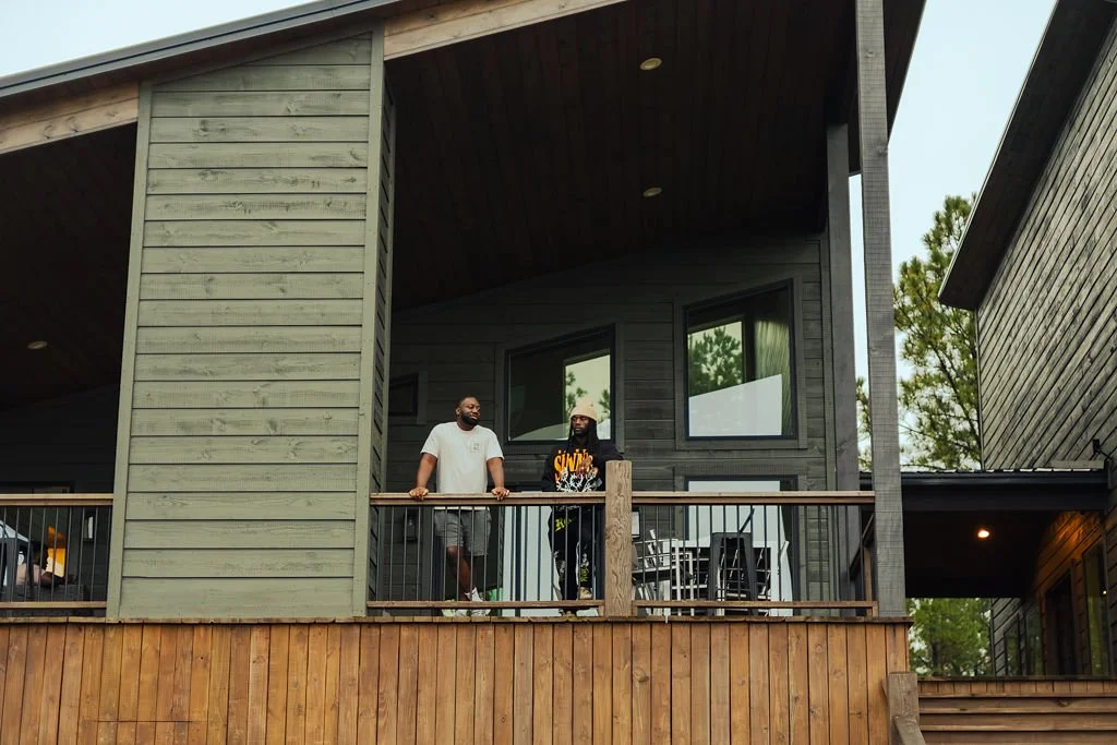 Two men standing on a balcony of a modern house with gray siding and large windows, surrounded by trees.